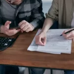 Двоє людей за столом заповнюють формуляр, поряд калькулятор, ручка, чашка - обговорють списання заборгованості за кредитом. Реструктуризація заборгованості