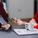 Дві бізнеследі в піджаках обговорюють оподаткування відчуження нерухомості, використовуючи калькулятор і вказуючи на мініатюрний будинок на контракті