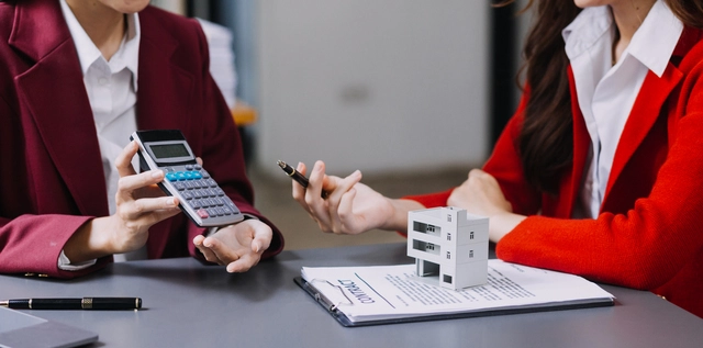 Дві бізнеследі в піджаках обговорюють оподаткування відчуження нерухомості, використовуючи калькулятор і вказуючи на мініатюрний будинок на контракті