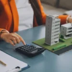 Жінка в помаранчевому піджаку біля моделей будівель на столі з блокнотом і ручкою обраховує на калькуляторі та у ноутбуці оподаткування продажу нерухомості